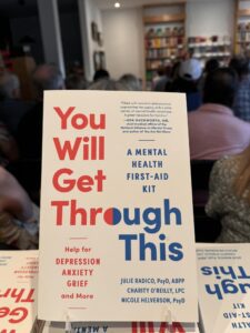 Picture of book cover "You Will Get Through This." Book signing attendees are seated in chairs in the background.