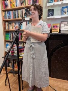 Charity O'Reilly speaking at a microphone. Book shelves are in the background.