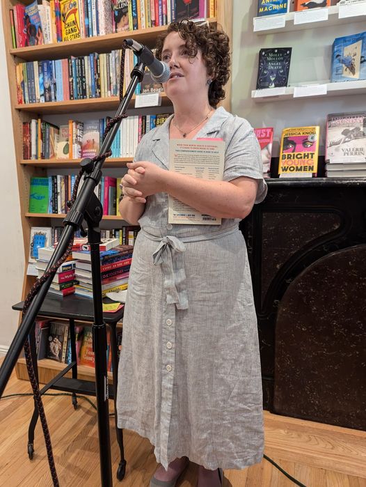 Charity O'Reilly speaking at a microphone. Book shelves are in the background.